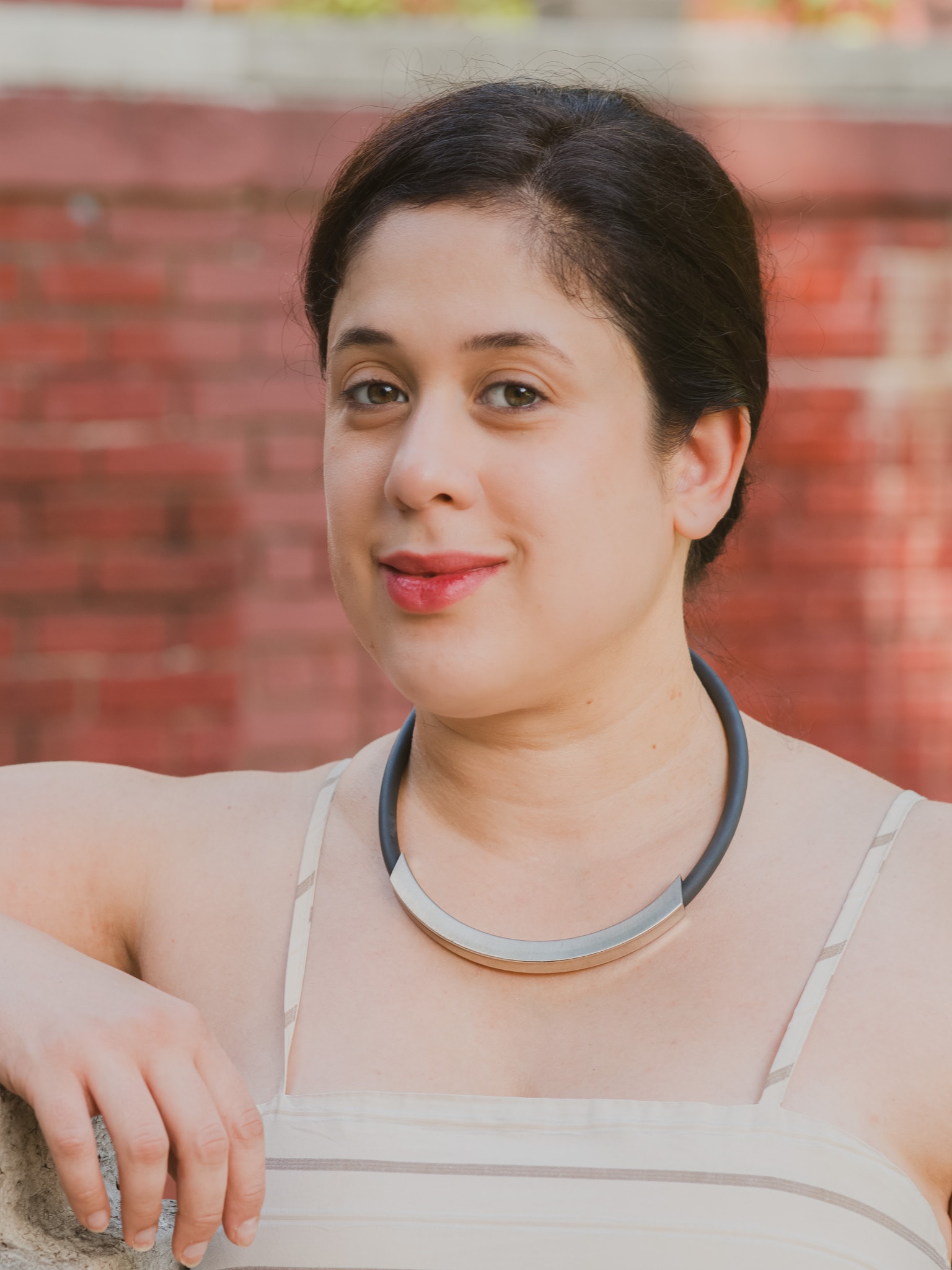Outdoor professional headshot of a woman wearing a modern silver statement necklace against a red brick background, by Fuentes Studio.