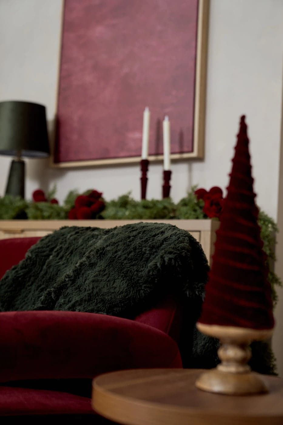 Taper candles and a pine garland with red roses on the sideboard, behind the green throw and a red velvet cone tree, in the Yonkers Christmas studio