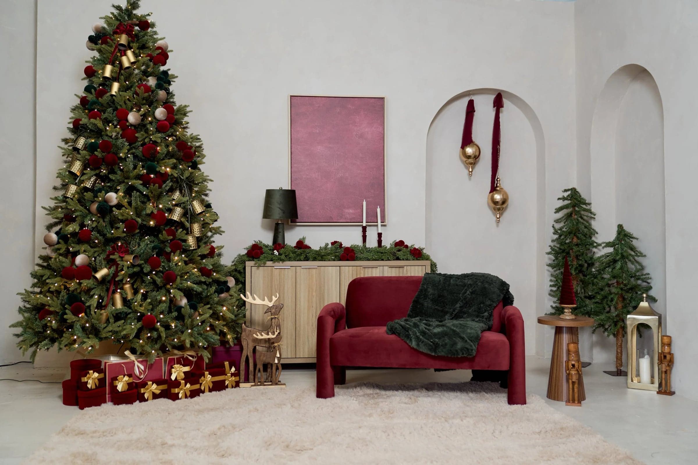 The Christmas studio set in Yonkers: a decorated tree with red and gold gifts, a wooden sideboard with a pine garland and candles, a burgundy velvet loveseat with a green throw, arched alcoves with gold ornaments and a white rug