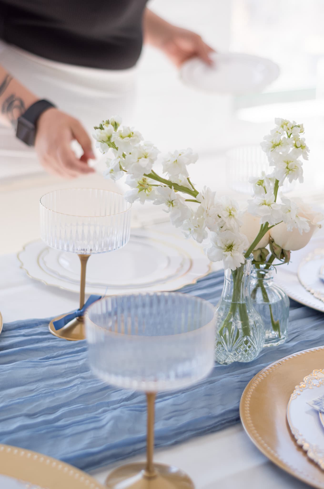 Hands arranging white stock flowers and ribbed glassware on the blue cheesecloth runner